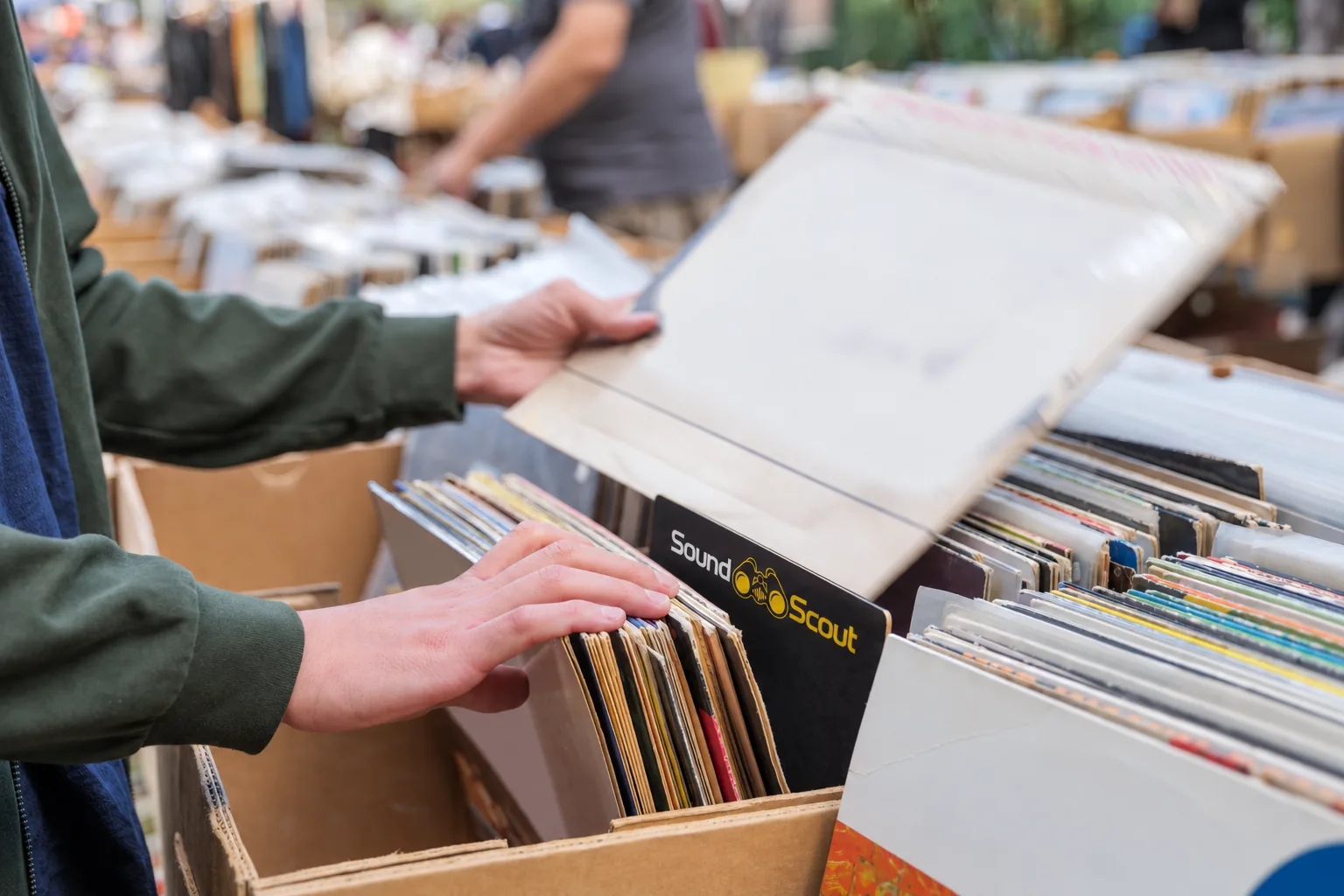 Person browsing through vinyl records at a record fair, with a subtle SoundScout-branded divider among the records.